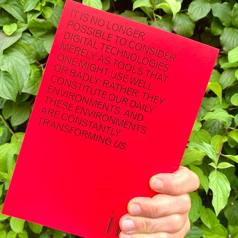 A hand holds a bright red book or booklet in front of green foliage. The cover features black text that reads, 'IT IS NO LONGER POSSIBLE TO CONSIDER DIGITAL TECHNOLOGIES MERELY AS TOOLS THAT ONE MIGHT USE WELL OR BADLY; RATHER, THEY CONSTITUTE OUR DAILY ENVIRONMENTS, AND THESE ENVIRONMENTS ARE CONSTANTLY TRANSFORMING US.