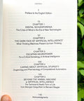 A hand holding an open book showing the table of contents page, with green foliage in the background.