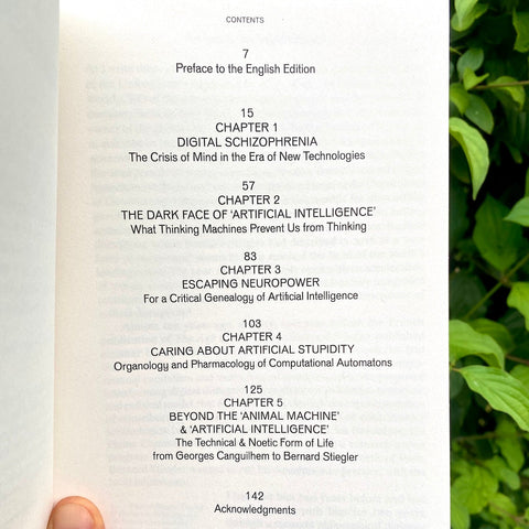 A hand holding an open book showing the table of contents page, with green foliage in the background.